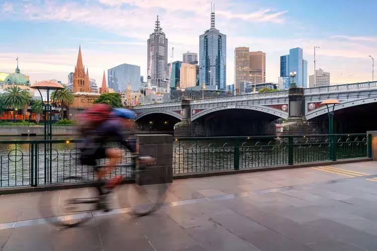 Cyclist riding beside a river with a city skyline behind